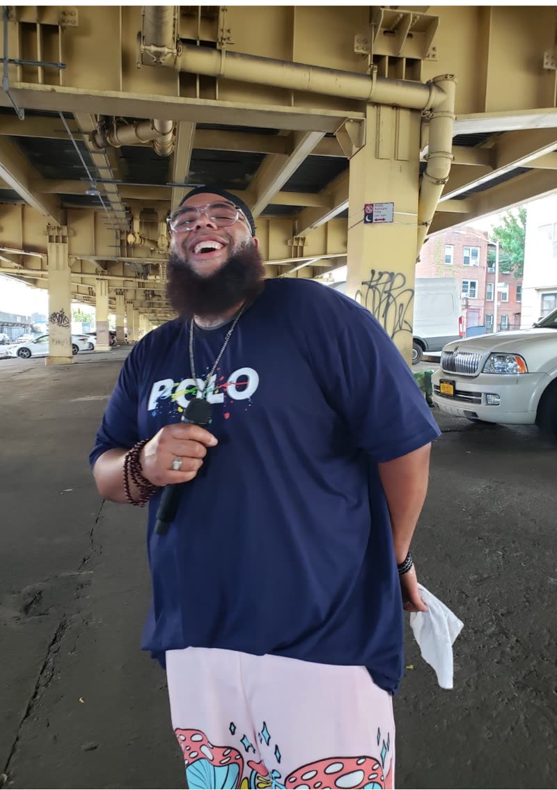 Barrz laughing with a handheld mic under the Brooklyn elevated tracks